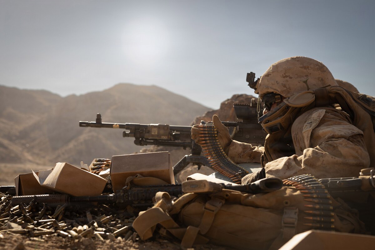 A man wearing a camouflage military uniform and a face mask holds a military rifle and a string of bullets in his hand; there are mountains in the background.
