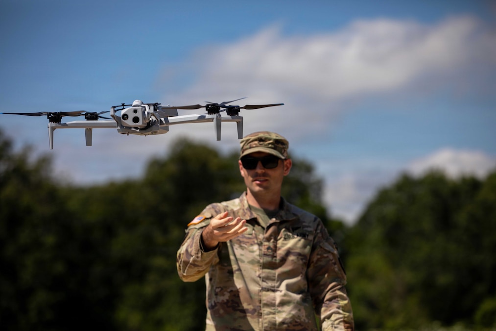 A soldier holds their hand out while a drone flies at eye level under a blue sky with a wooded area in the background.