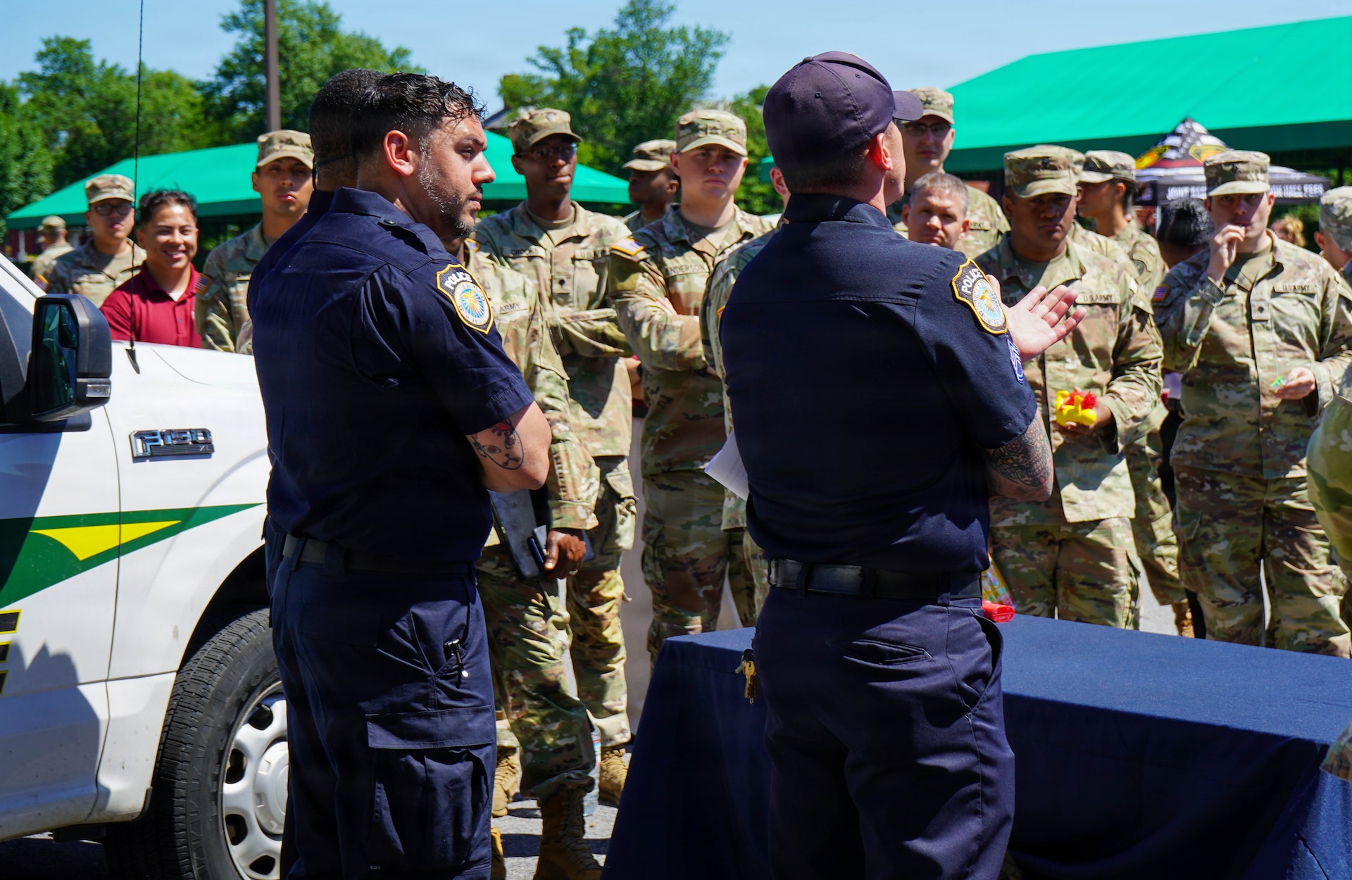 Two people wearing dark blue utility uniforms are facing a large group of people, mostly military members in combat uniforms, during an outdoor event. There is a service truck to the left and green awnings are visible in the background