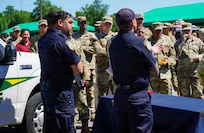 Two people wearing dark blue utility uniforms are facing a large group of people, mostly military members in combat uniforms, during an outdoor event. There is a service truck to the left and green awnings are visible in the background
