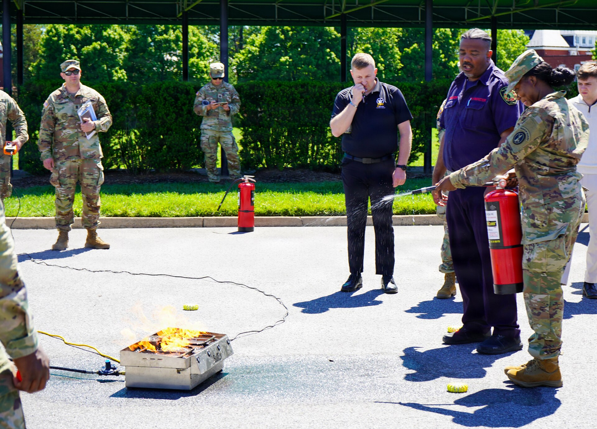 An Army soldier in combat uniform is putting out a fire with a fire extinguisher while other people watch from a safe distance.
