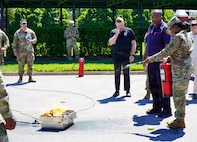 An Army soldier in combat uniform is putting out a fire with a fire extinguisher while other people watch from a safe distance.