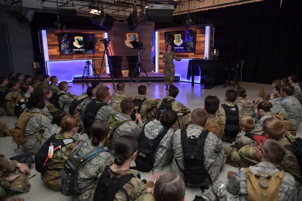 A woman in military uniform speaks to a group of civil air patrol cadets as they sit on a floor inside of a tv recording studio.