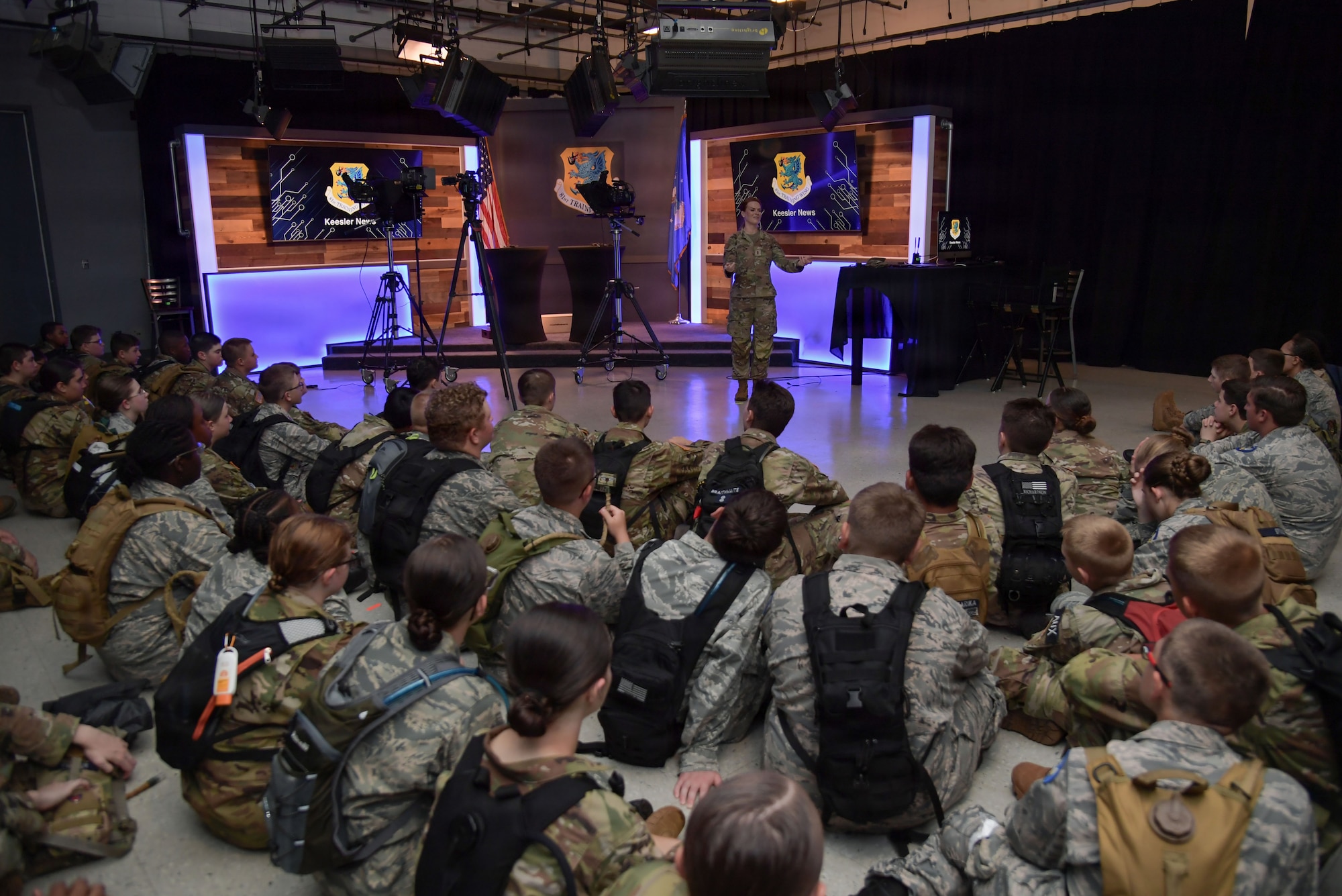 A woman in military uniform speaks to a group of civil air patrol cadets as they sit on a floor inside of a tv recording studio.