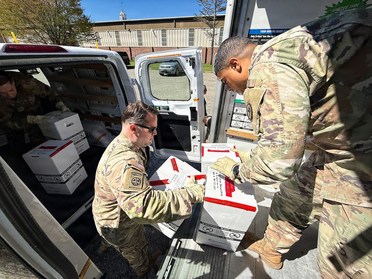 Two men wearing camouflage military uniforms move boxes from a van into a truck where another man in similar attire checks them.