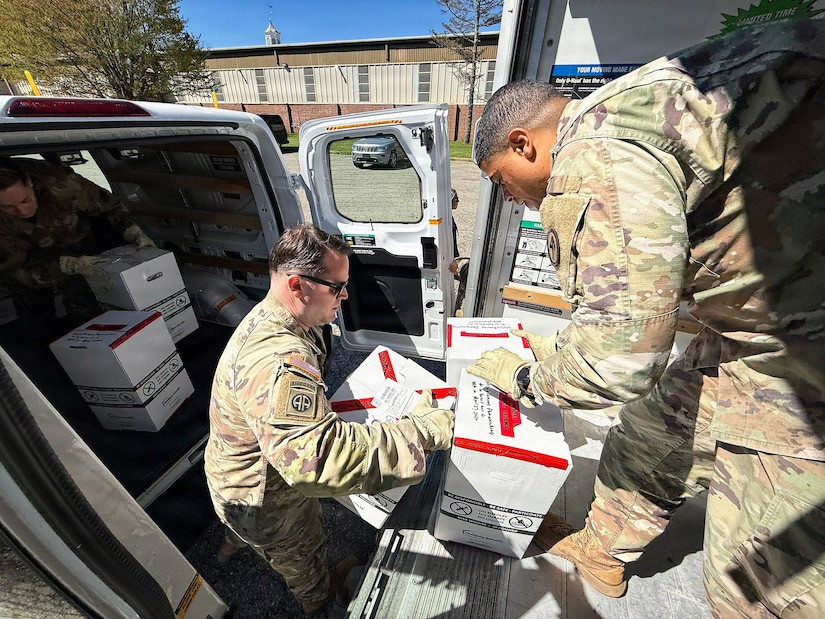 Two men wearing camouflage military uniforms move boxes from a van into a truck where another man in similar attire checks them.