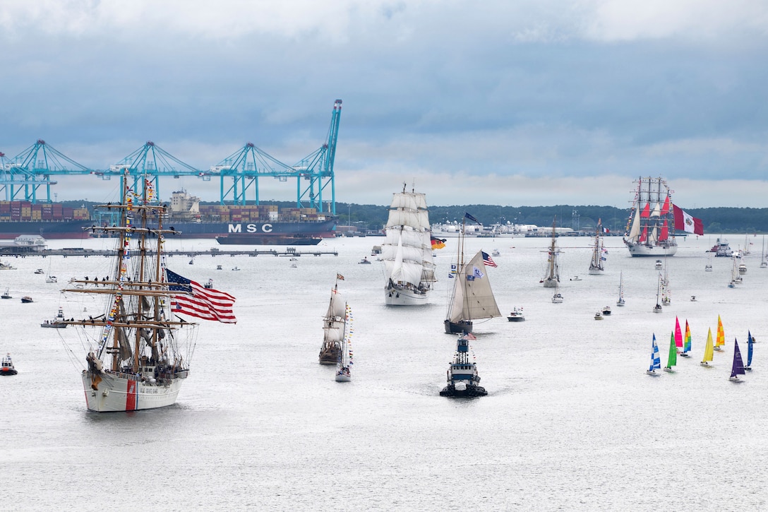 Sailboats and other types of vessels, some with American and service flags, are in the water near a port. Cargo ships and equipment are in the background.