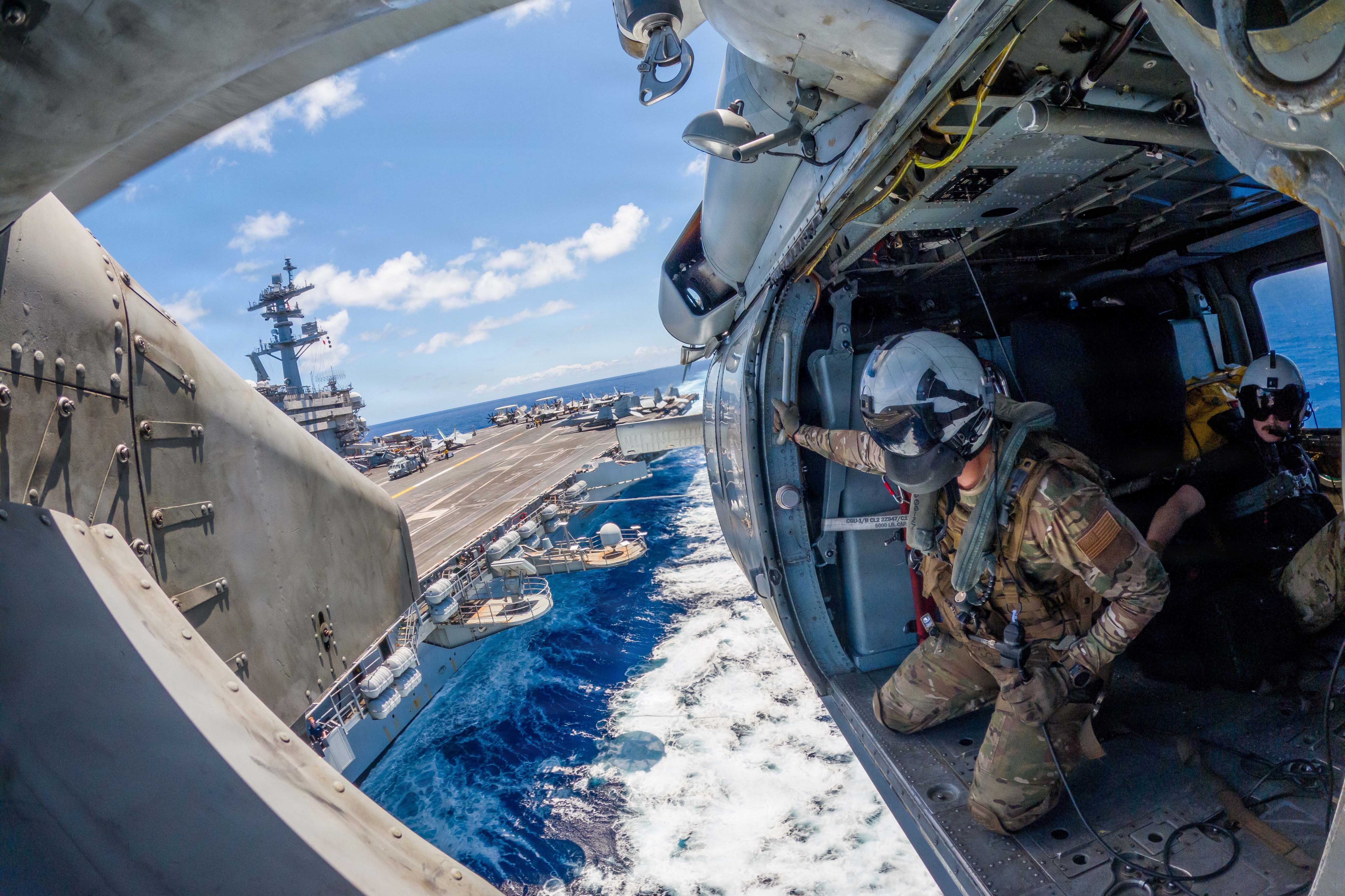 A sailor looks out over blue water from the open door of a helicopter flying near a ship at sea.