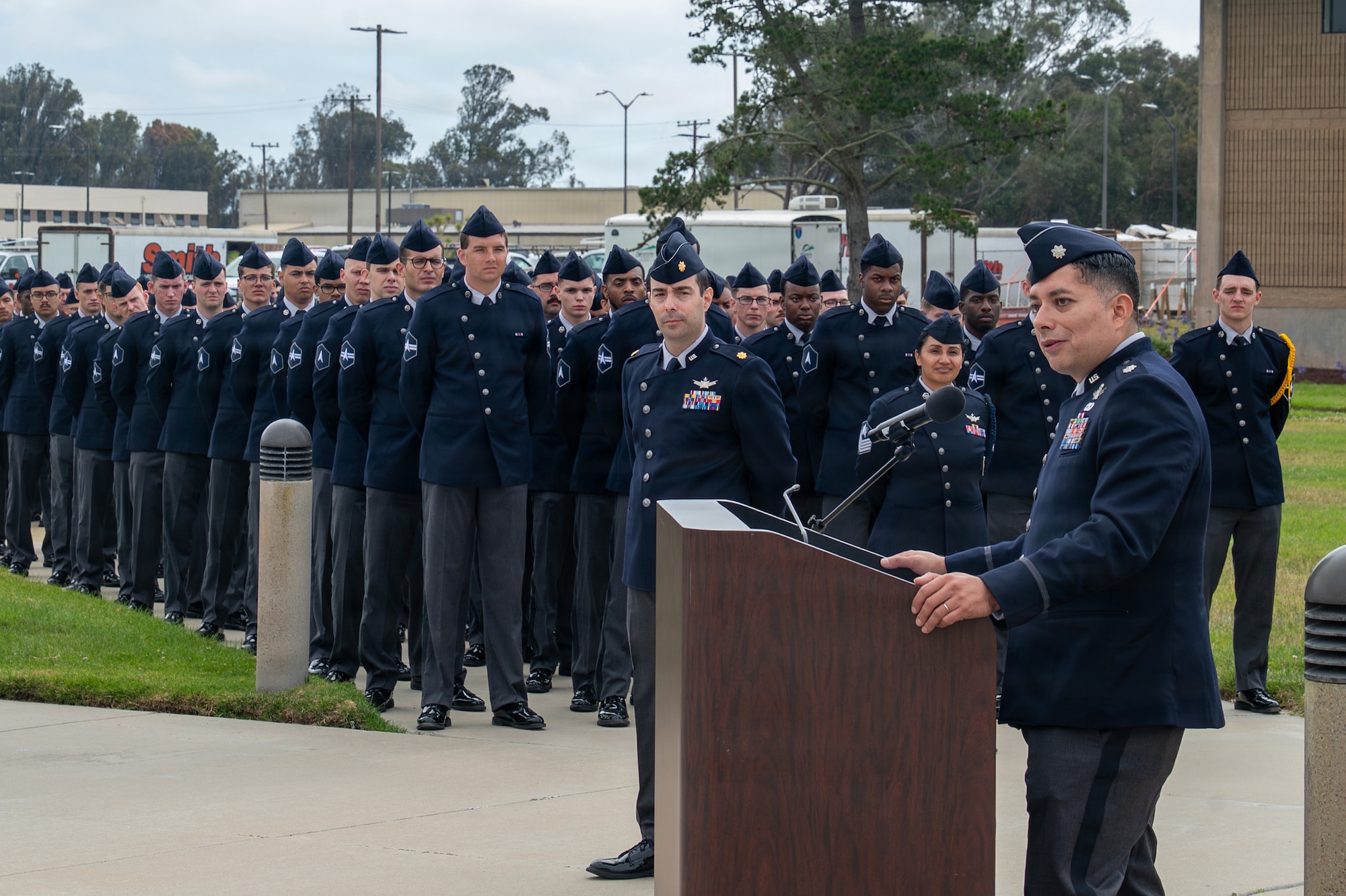 A person stands behind a podium and speaks while many people stand at ease and listen in formation