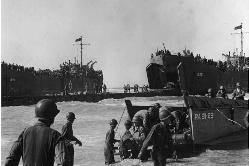 Troops in landing boats disembark and move through shallow water toward a beach.