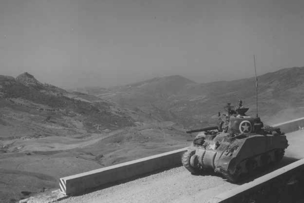 A military tank crosses a bridge with a valley and mountains in the background.