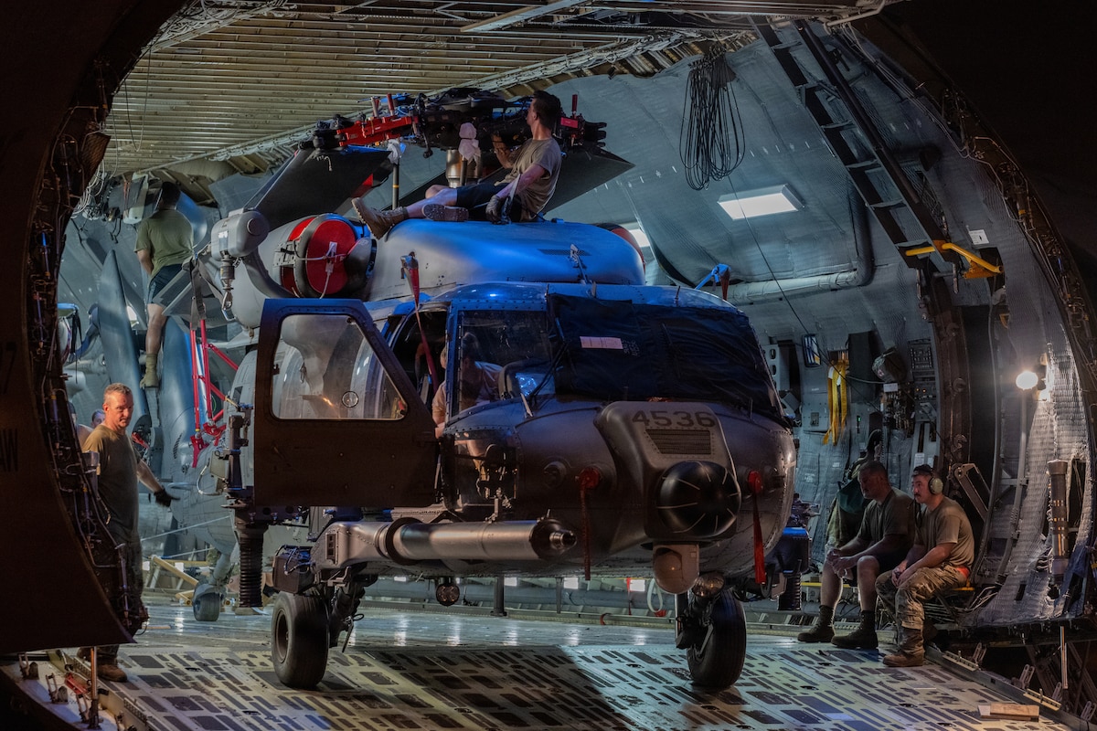 Eight people wearing military uniforms and casual attire maneuver a military helicopter at night that is inside the cargo bay of a larger military aircraft.