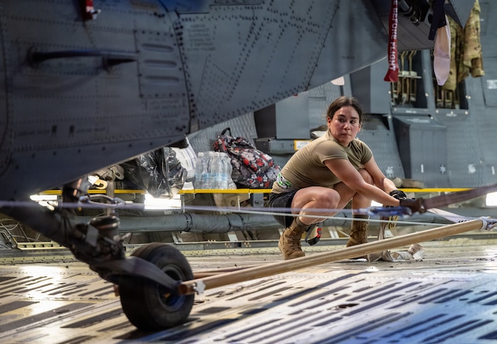 A woman wearing casual attire pulls on a strap to maneuver a military helicopter that is inside the cargo bay of a larger military aircraft.