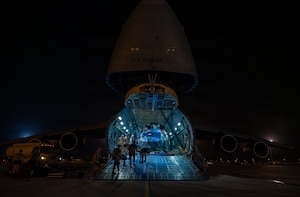 A dozen people wearing military uniforms and casual attire maneuver a military helicopter at night that is inside the cargo bay of a larger military aircraft.