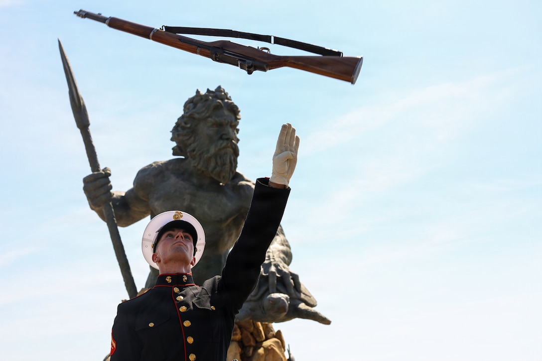 A Marine in ceremonial dress tosses a rifle toward a blue sky in front of a statue.