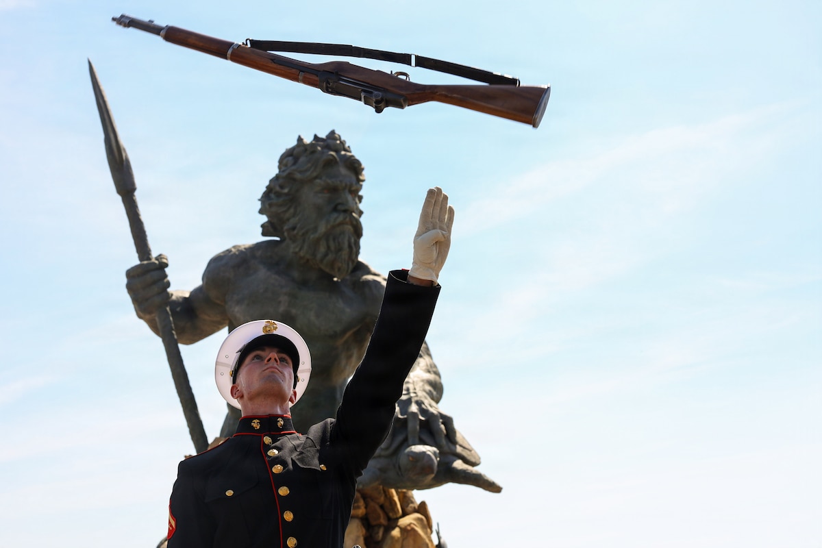 A Marine in ceremonial dress tosses a rifle toward a blue sky in front of a statue.