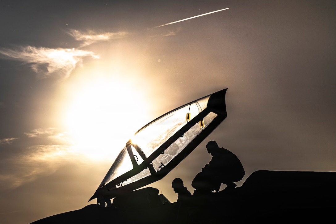 An airman sits an open cockpit of an aircraft while a fellow airman kneels behind as seen in silhouette under a sunlit sky.