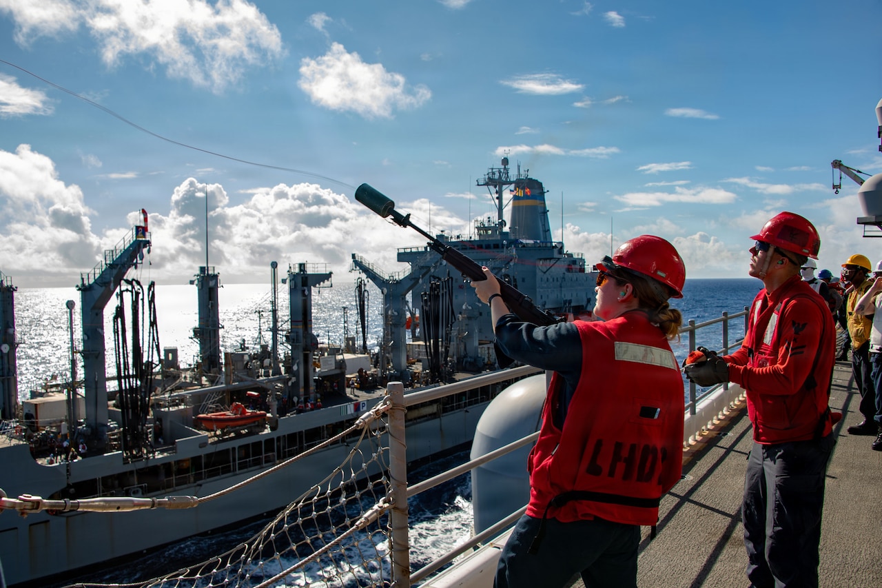 A sailor in a red vest and hard hat aims a gun from the deck of one ship toward another, as a fellow sailor stands by.