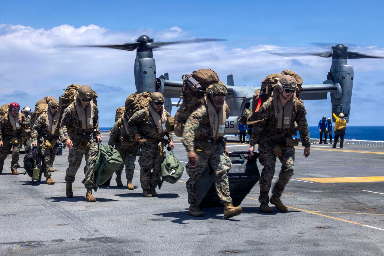 Marines carry luggage on a ship's deck near a parked aircraft.
