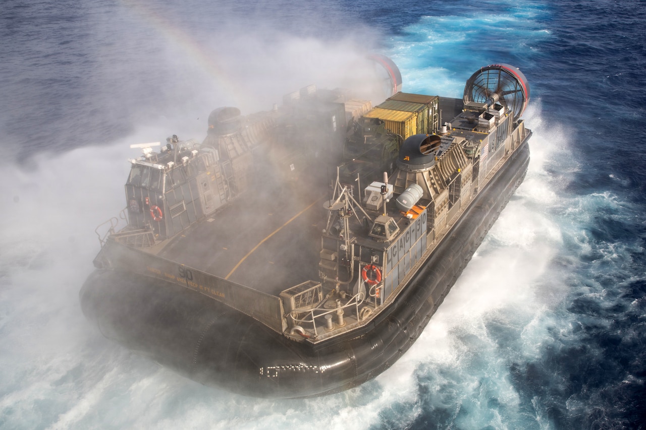 An air-cushioned landing craft splashes in blue water, creating a white wake and sending mist into the air.