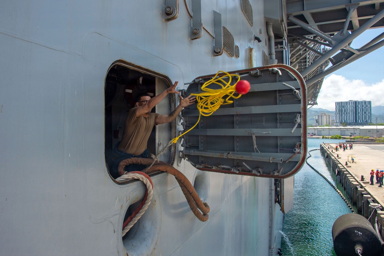 A sailor tosses a yellow line from an open hatch on the side of a ship toward a pier.