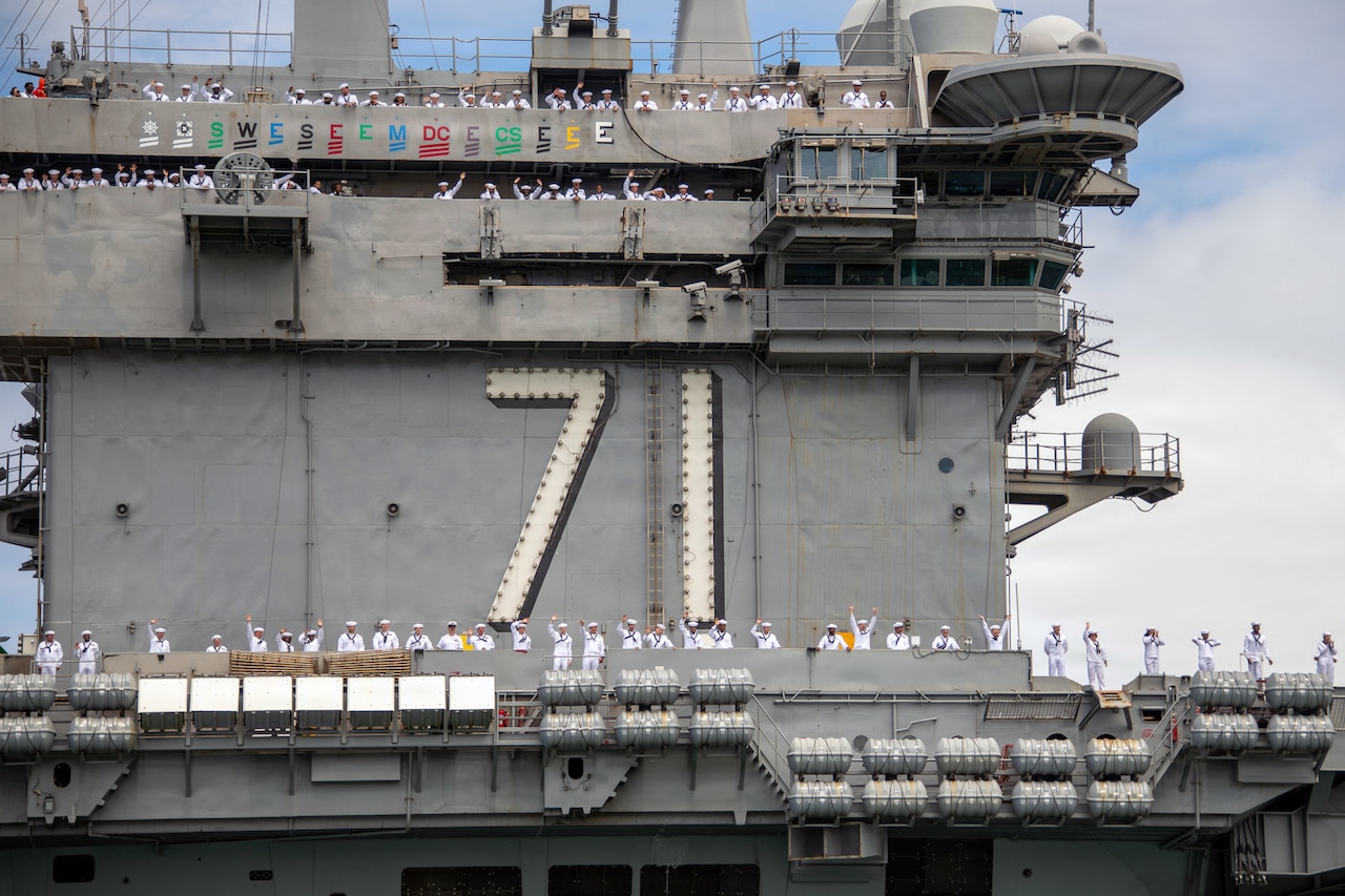 Sailors in white uniforms wave while manning the rails of an aircraft carrier with the hull number "71" on its side.