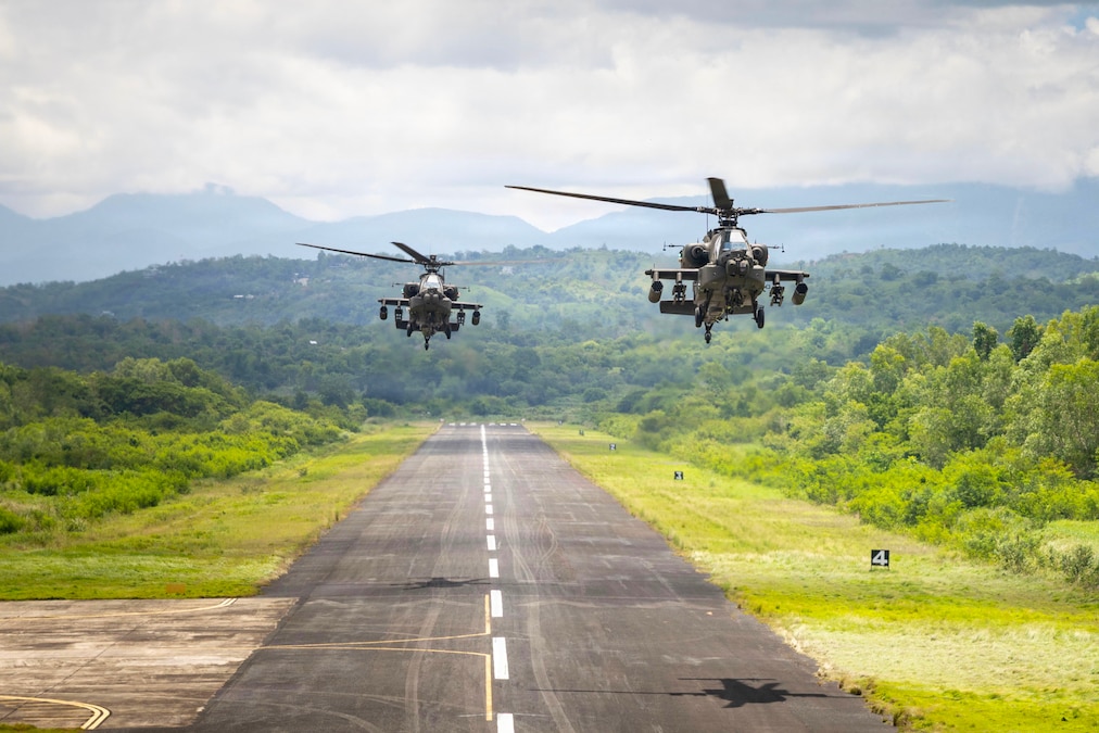 Two helicopters hover over a runway that has a dashed line down the center. Green trees and mountains are in the background.