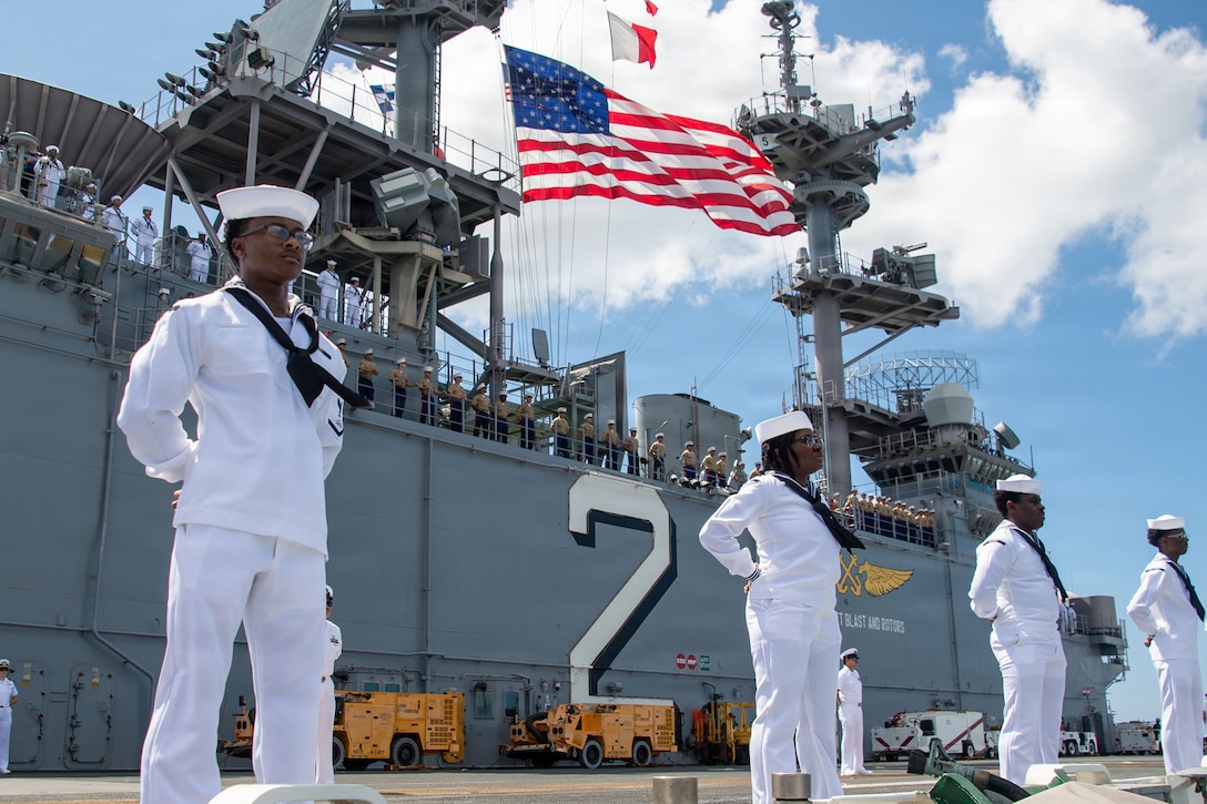 Four sailors stand at the rim of a ship's deck, holding their arms behind their backs. Other sailors at the rail of the upper deck and a U.S. flag are in the background.