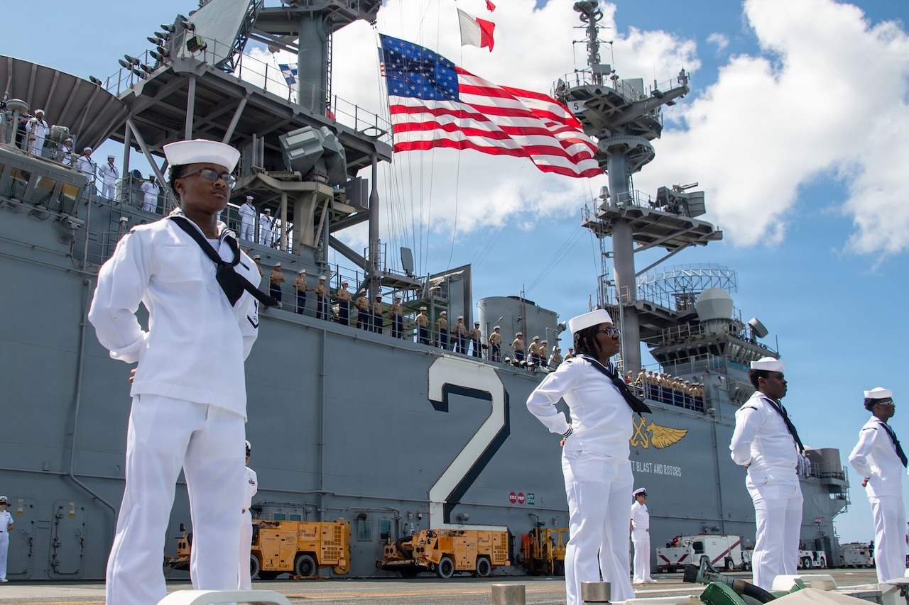 Four sailors stand at the rim of a ship's deck, holding their arms behind their backs. Other sailors at the rail of the upper deck and a U.S. flag are in the background.