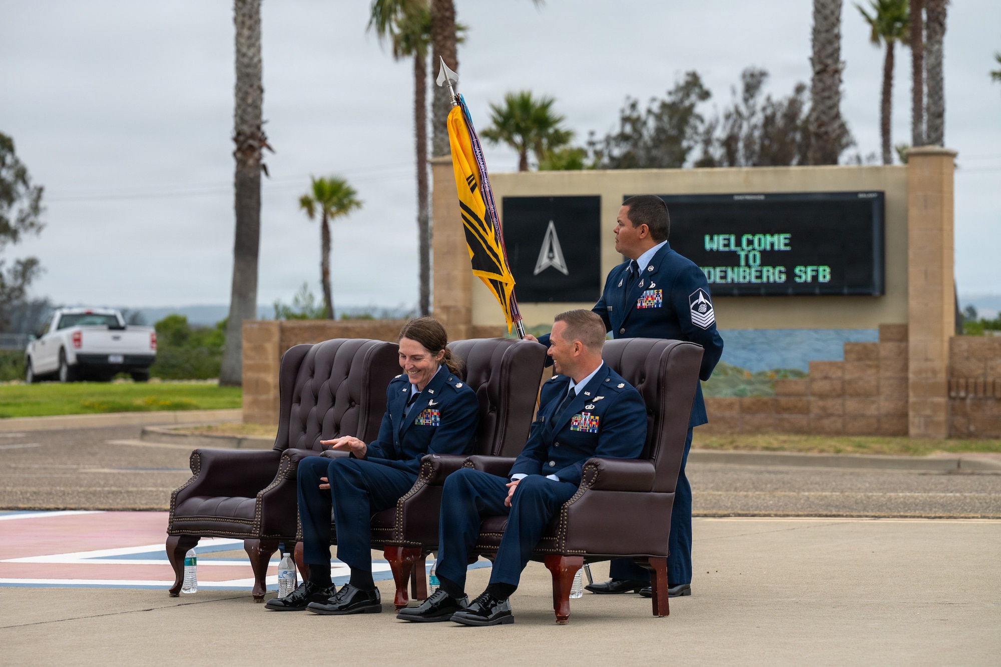 The incoming and outgoing 30th Operations Support Squadron commanders sit on leather chairs at the front of the change of command ceremony and the first sergeant stands behind them holding the guidon.