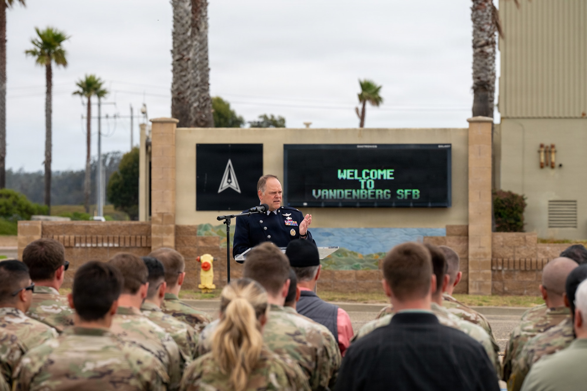 On an airfield with palm trees in the background the commander of Vandenberg Space Force Base gives a speech from a podium in front of a crowd.