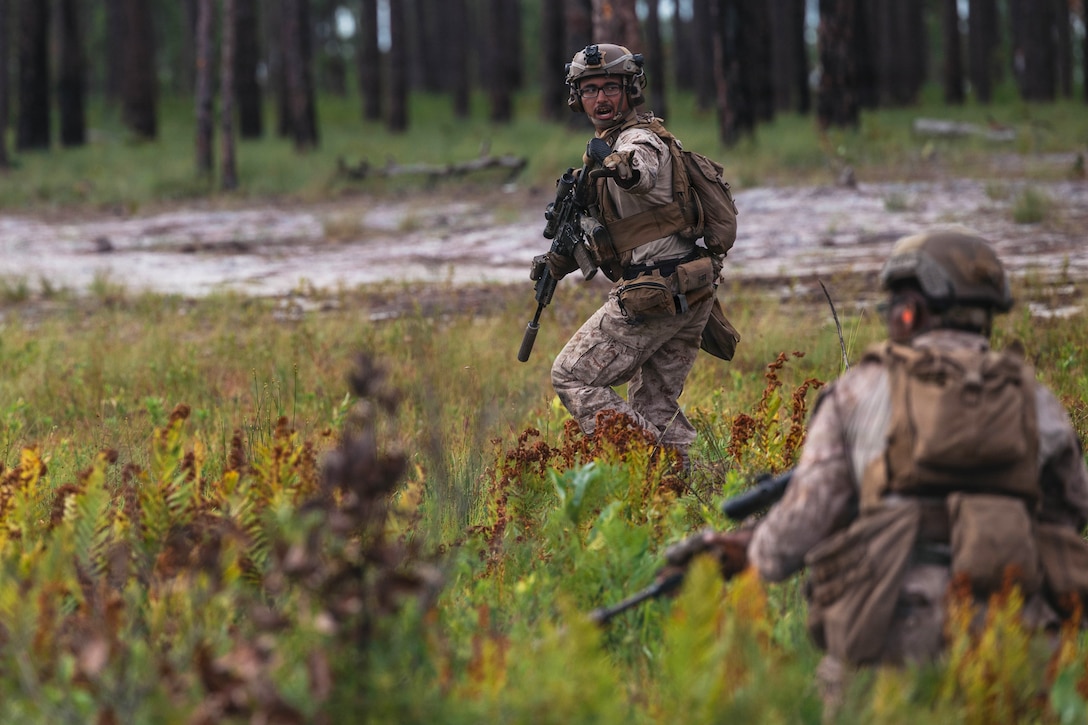 A Marine holding a rifle looks back and motions to another Marine to stay put, while in a field with tall weeds and trees in the background.