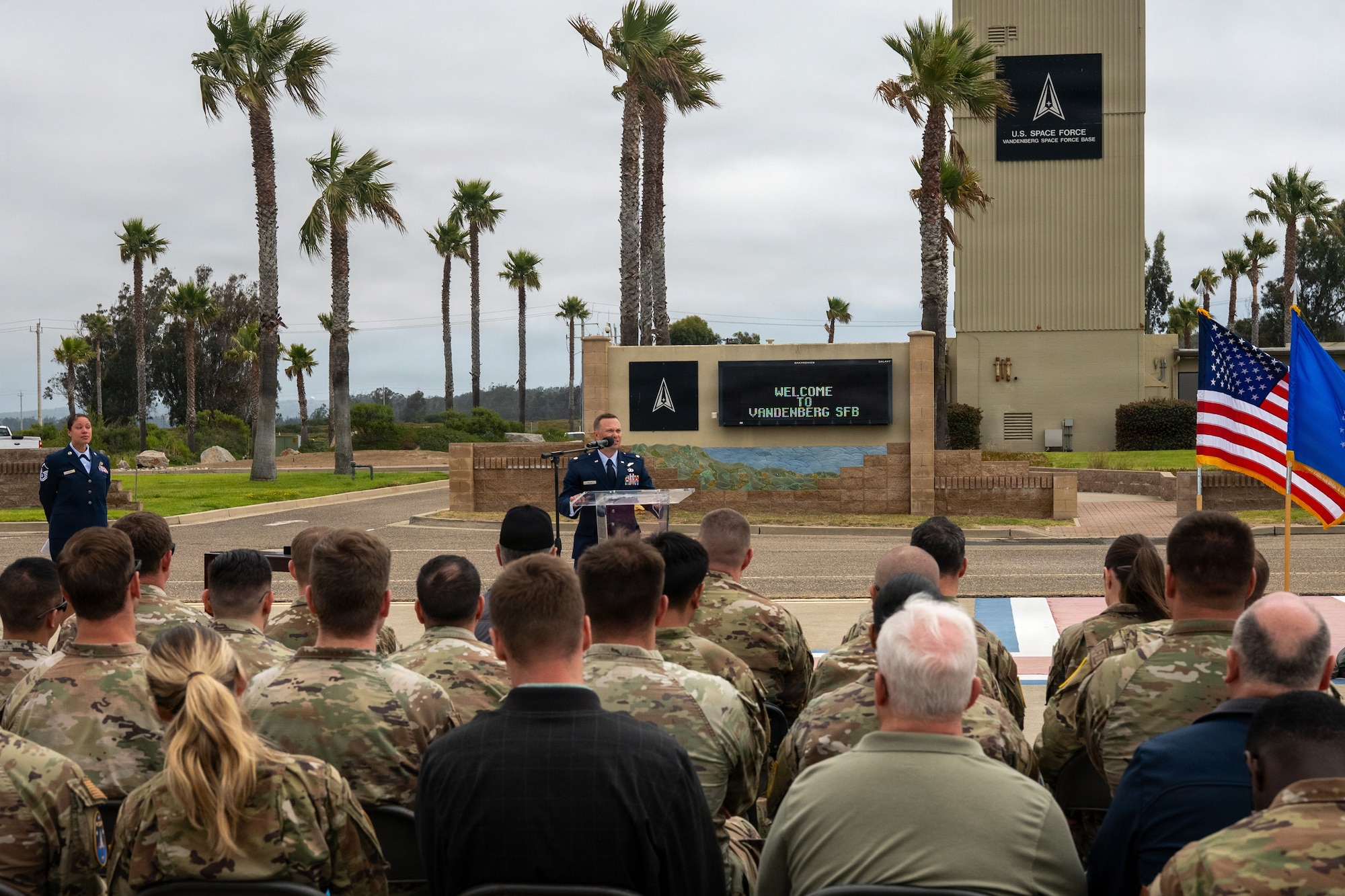 On an airfield with palm trees in the background the incoming squadron commander speaks to a crowd of military members, friend and family.