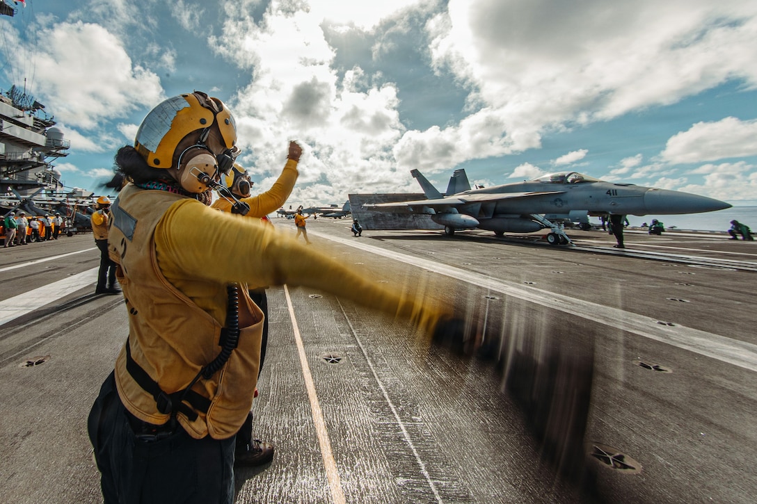 A sailor in yellow attire directs a fighter jet on the deck of a military ship.