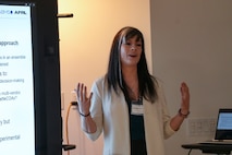a woman stands in between two screens speaking