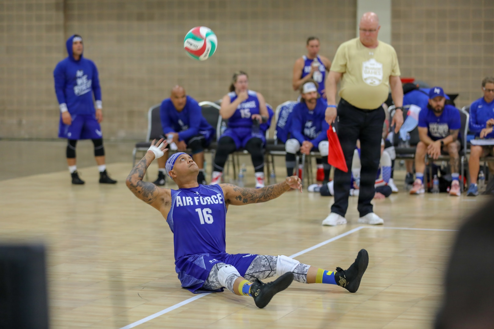 Air Force Warrior is seated on the basketball court and tosses the ball up in the air as he prepares to serve. A line judge is standing next to him watching him serve.
