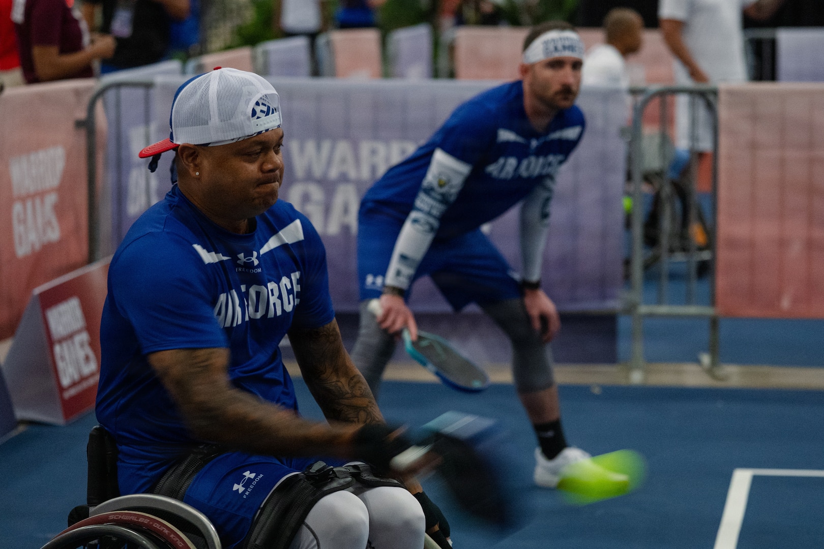 Two athletes are on a pickle ball court. One athlete is in a wheelchair holding his paddle and the other is standing holding his paddle.