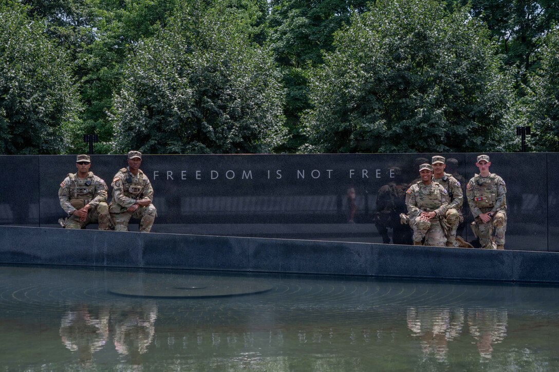 Soldiers pose in front of a black wall inscribed with "freedom is not free" outside, with a shallow reflecting pool in front of them.