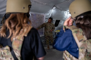 A service member points at a map in front of two individuals in full combat gear.