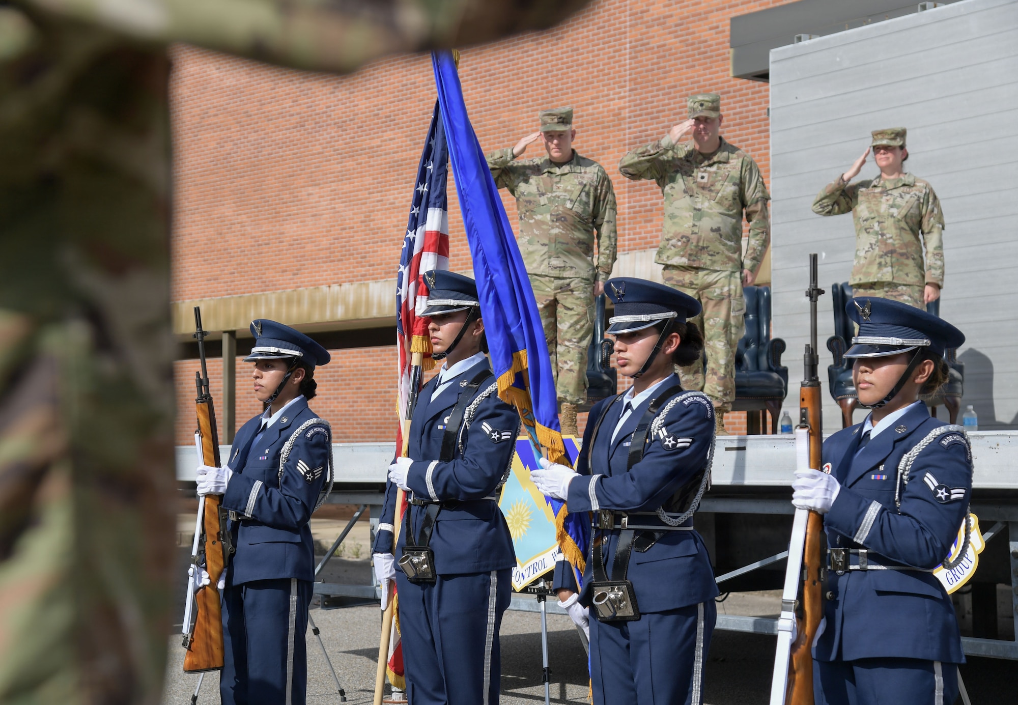 A group of honor guard members present the U.S. and Air Force flags while military members render a salute in the background from a stage.