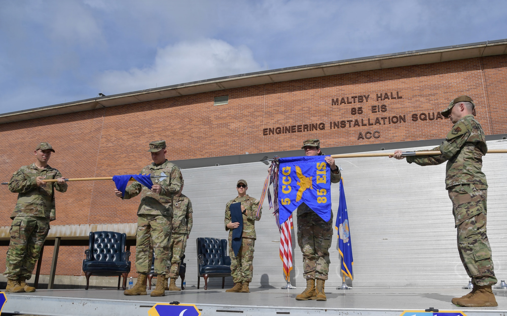 People in military uniforms stand on stage as they unfurl and furl guidons.