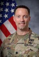 An Army Soldier wearing combat uniform seated in front of the U.S. flag for an official photo.