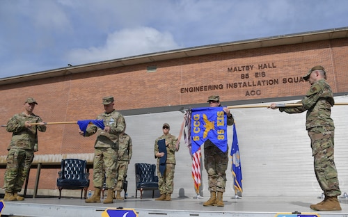 People in military uniforms stand on stage as they unfurl and furl guidons.