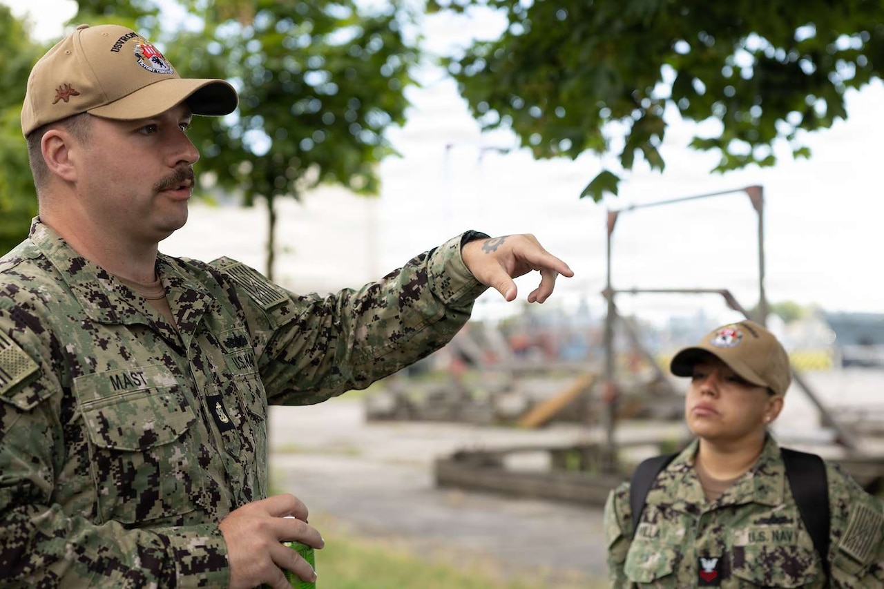 A man wearing a camouflage military uniform points to something in front of him as he speaks. Another person in similar attire watches.