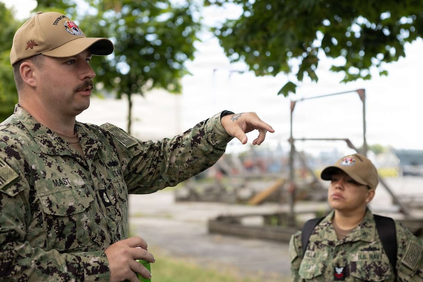 A man wearing a camouflage military uniform points to something in front of him as he speaks. Another person in similar attire watches.