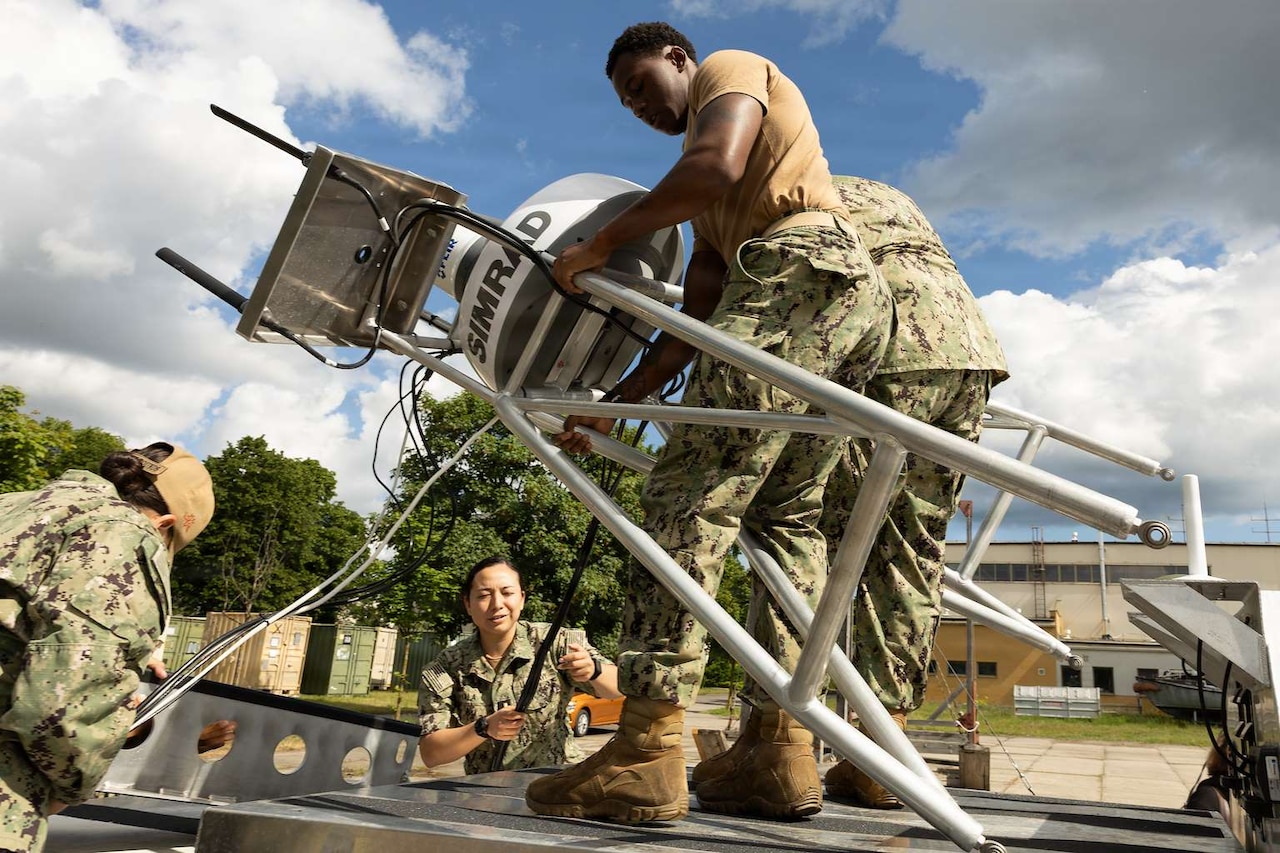 People wearing camouflage military uniforms work on a small military unmanned surface vessel. Two of them hold up one of its parts at an angle while two others unravel cords attached to the craft.
