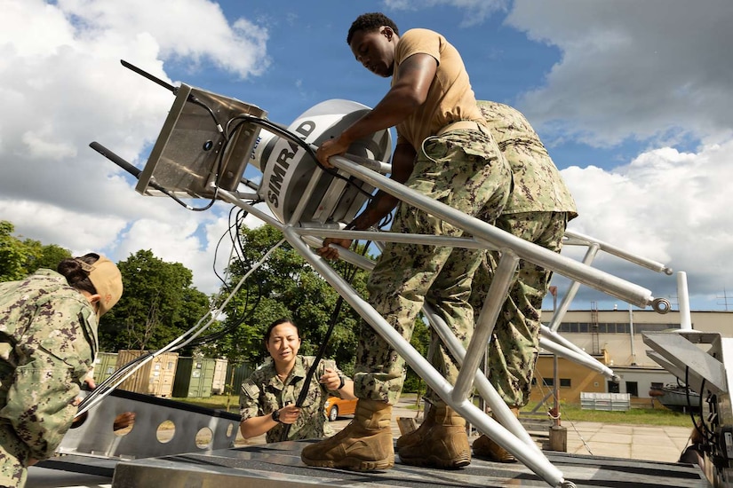 People wearing camouflage military uniforms work on a small military unmanned surface vessel. Two of them hold up one of its parts at an angle while two others unravel cords attached to the craft.