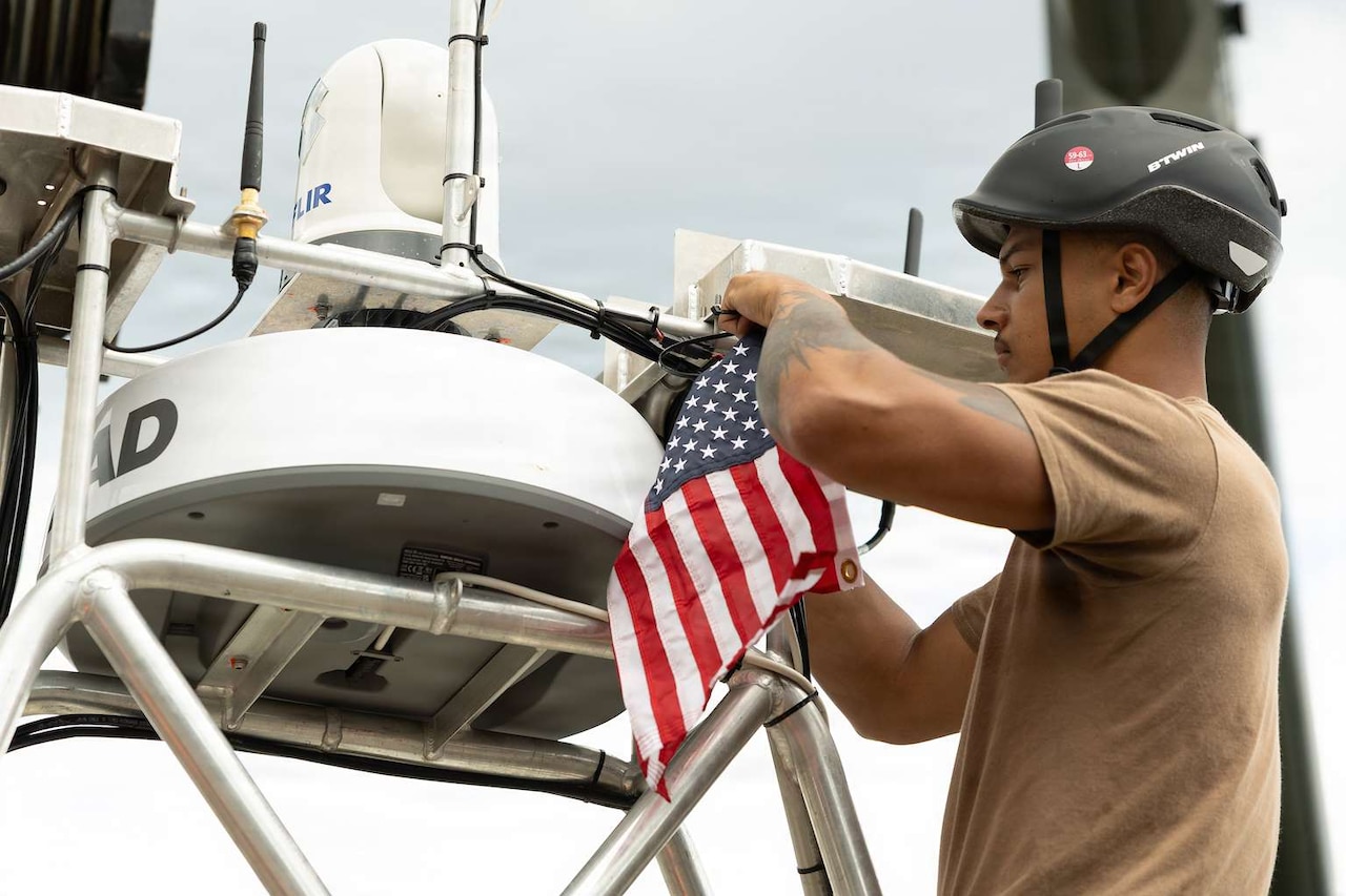 A man wearing a brown T-shirt and helmet, hangs a small American flag onto an unmanned surface vessel.