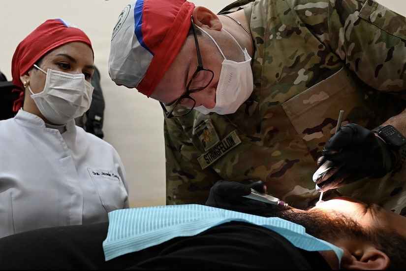 A man in a camouflage military uniform, medical hat, face mask and gloves checks the teeth of a man lying down in a dental chair as another woman in medical attire observes.