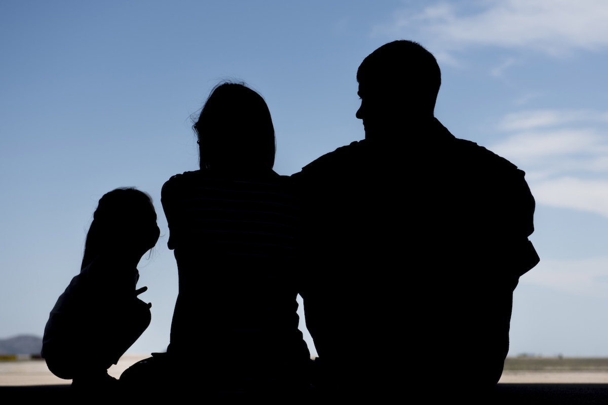 A silhouette of two adults and a child sitting together facing a blue sky.